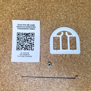 May include: A white semi-circular plastic component with three rectangular cutouts, a small metal nut, and a thin metal rod. A white paper card is included with a QR code and the text: Scan the QR code below to watch the installation video. Or visit the KIN Additive YouTube Channel.