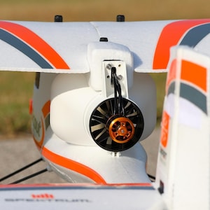 May include: Close-up of a white and orange model airplane. The plane features a black propeller with an orange center and a white body. The wings have orange and gray accents. The model is designed for remote control.