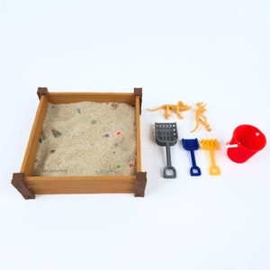 May include: A wooden sandbox with a brown frame and sand inside. There are small plastic gems and seashells scattered in the sand.  There are two plastic dinosaur toys, a gray plastic shovel, a blue plastic shovel, a yellow plastic rake, and a red plastic bucket.