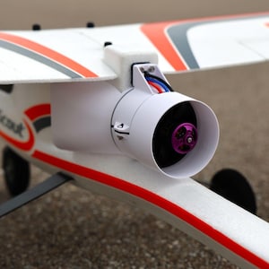 May include: A white and red model airplane with a jet engine. The plane has a white fuselage and wings with red and gray accents. The jet engine is white with a purple propeller.
