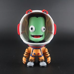 Peut inclure: Une figurine d'astronaute extraterrestre verte portant un casque blanc et rouge avec un grand sourire. La figurine porte une combinaison spatiale orange et bleue.