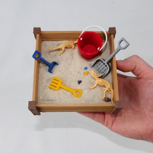 May include: A miniature wooden sandbox with a red bucket, a gray shovel, a yellow rake, and two small dinosaur skeletons. The sandbox is sitting on a hand.