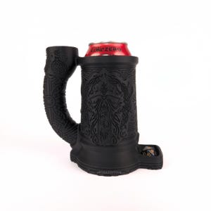 May include: Black silicone mug with a detailed design of a face and intricate patterns. The mug has a handle and a built-in dice tray. A can of Coke Zero is in the mug.