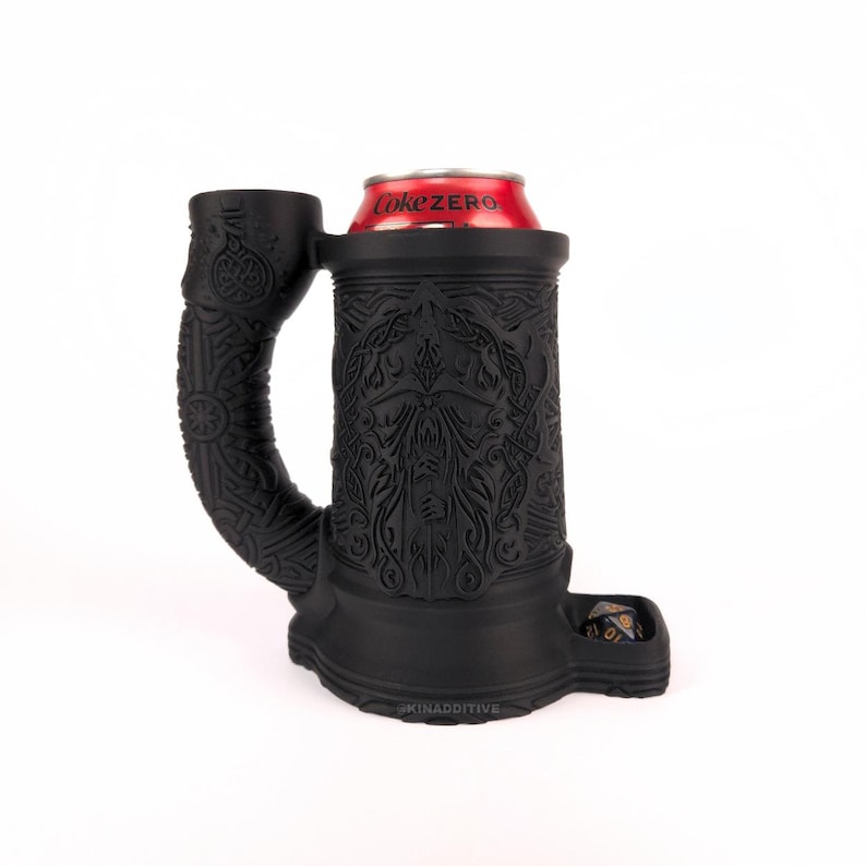 May include: Black silicone mug with a detailed design of a face and intricate patterns. The mug has a handle and a built-in dice tray. A can of Coke Zero is in the mug.