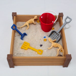 May include: A wooden sandbox with a red bucket, a blue shovel, a yellow rake, and two beige dinosaur skeletons. There are also small, colorful gems scattered in the sand.