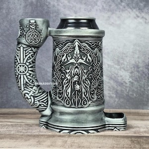 May include: A silver metal beer stein with a detailed design of a bearded figure with a sword. The stein has a handle with a Celtic knot design.