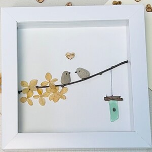 May include: A white framed picture with a white background. The picture features two small birds made of stone perched on a branch with dried flowers. A small birdhouse made of green glass hangs from the branch. The word "Love" is written on a small wooden heart in the upper right corner of the picture.