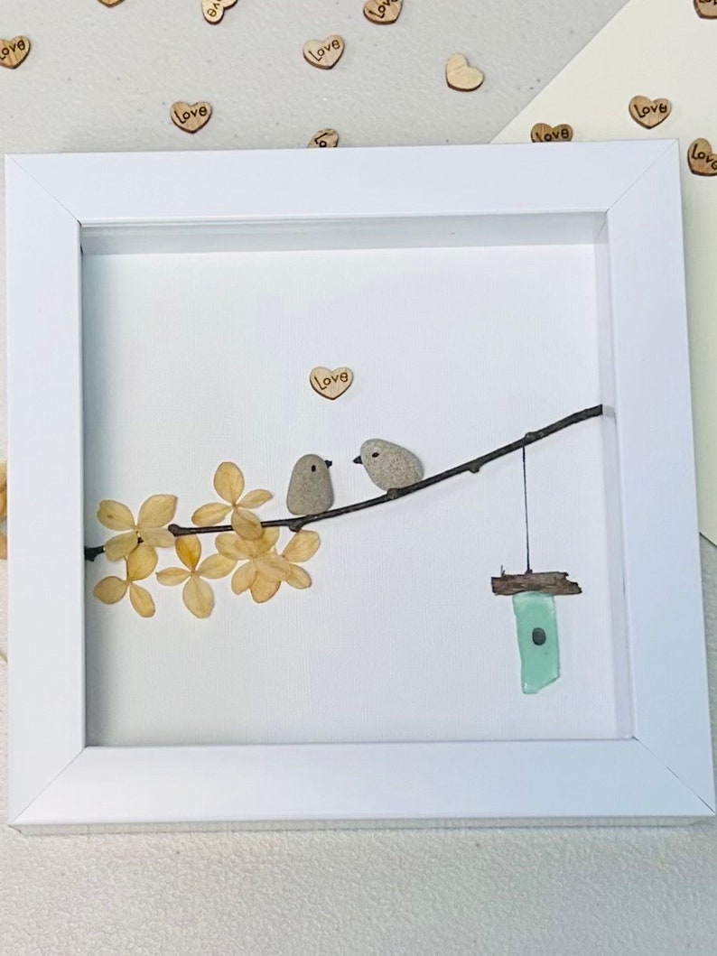 May include: A white framed picture with a white background. The picture features two small birds made of stone perched on a branch with dried flowers. A small birdhouse made of green glass hangs from the branch. The word "Love" is written on a small wooden heart in the upper right corner of the picture.