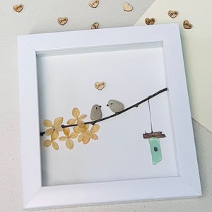 May include: A white framed picture with a white background. Two small grey birds are perched on a branch with dried flowers. A small blue glass birdhouse hangs from the branch. The word "Love" is written on a small wooden heart above the birds.