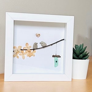 May include: A framed artwork featuring two small grey birds perched on a twig with a small wooden birdhouse hanging below. The birds are surrounded by dried white flowers and a small heart with the word "love" written on it.