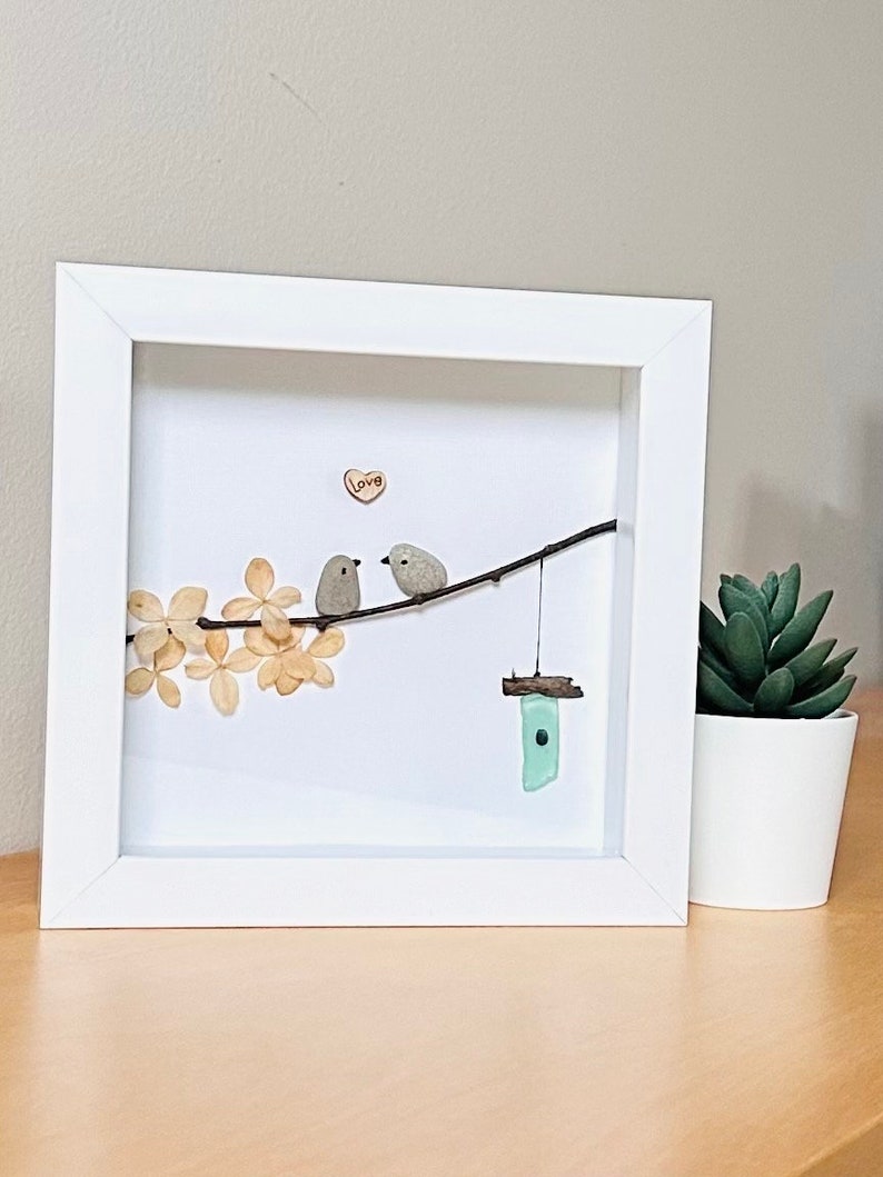May include: A framed artwork featuring two small grey birds perched on a twig with a small wooden birdhouse hanging below. The birds are surrounded by dried white flowers and a small heart with the word "love" written on it.