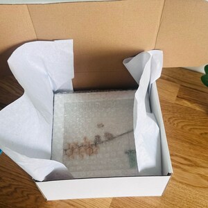 May include: A white cardboard box with a brown cardboard lid. The box is filled with white tissue paper and contains a framed picture. The picture is of a floral design with a white background.