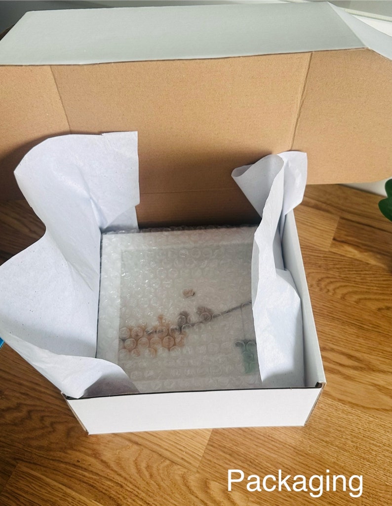 May include: A white cardboard box with a brown cardboard lid. The box is filled with white tissue paper and contains a framed picture. The picture is of a floral design with a white background.