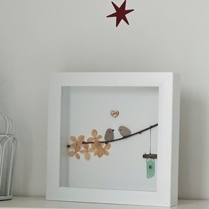 May include: A framed artwork featuring two small grey birds perched on a branch with brown leaves. The birds are facing each other and a small wooden heart with the word "loving" is above them. A small blue and green glass wind chime hangs from the branch.