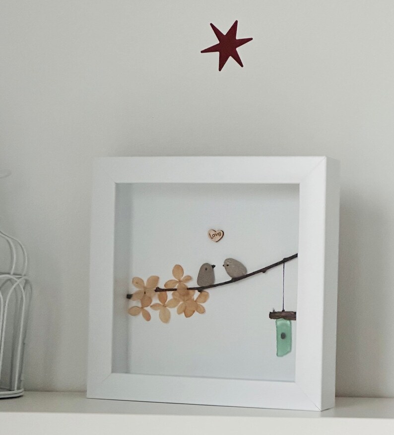 May include: A framed artwork featuring two small grey birds perched on a branch with brown leaves. The birds are facing each other and a small wooden heart with the word "loving" is above them. A small blue and green glass wind chime hangs from the branch.