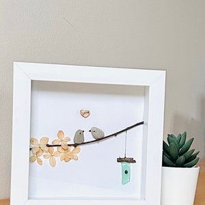 May include: Framed artwork featuring two small grey birds perched on a twig with a heart that says "Love" above them. The birds are made of painted stones and the twig is surrounded by dried white flowers. A small blue glass birdhouse hangs from the twig.
