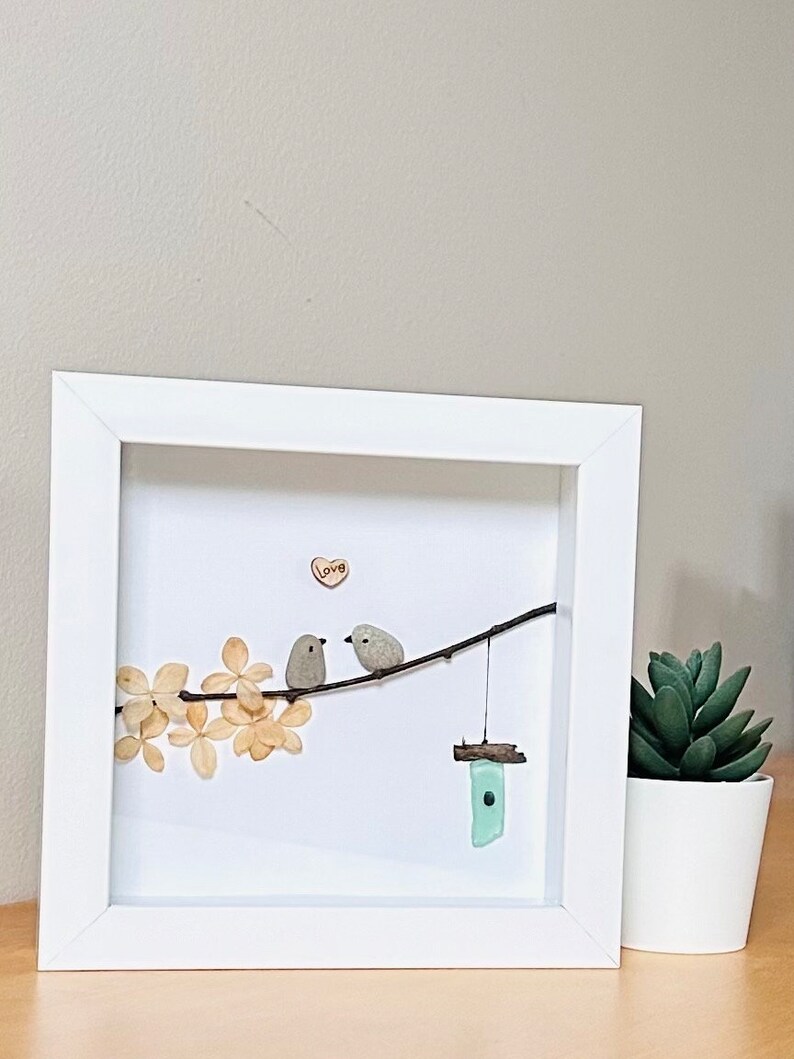 May include: Framed artwork featuring two small grey birds perched on a twig with a heart that says "Love" above them. The birds are made of painted stones and the twig is surrounded by dried white flowers. A small blue glass birdhouse hangs from the twig.