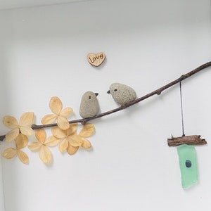 May include: A framed artwork featuring two small grey birds perched on a twig with a wooden heart that says "Love" above them. The twig is surrounded by dried beige flowers and a small green glass birdhouse hangs from the twig.