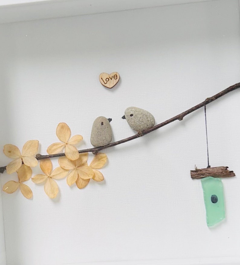 May include: A framed artwork featuring two small grey birds perched on a twig with a wooden heart that says "Love" above them. The twig is surrounded by dried beige flowers and a small green glass birdhouse hangs from the twig.