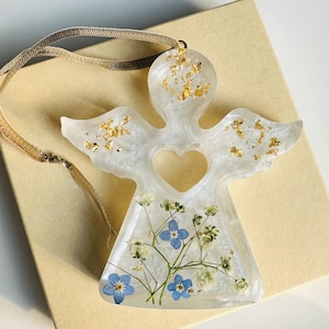 May include: A clear resin angel ornament with gold leaf inclusions and a heart-shaped void. The angel's wings and body are decorated with small blue and white flowers. A beige ribbon is attached for hanging. The ornament rests on a beige box.