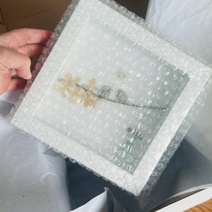 May include: A white frame with a clear plastic cover, protecting a delicate pressed flower arrangement. The frame is surrounded by bubble wrap for safe shipping.