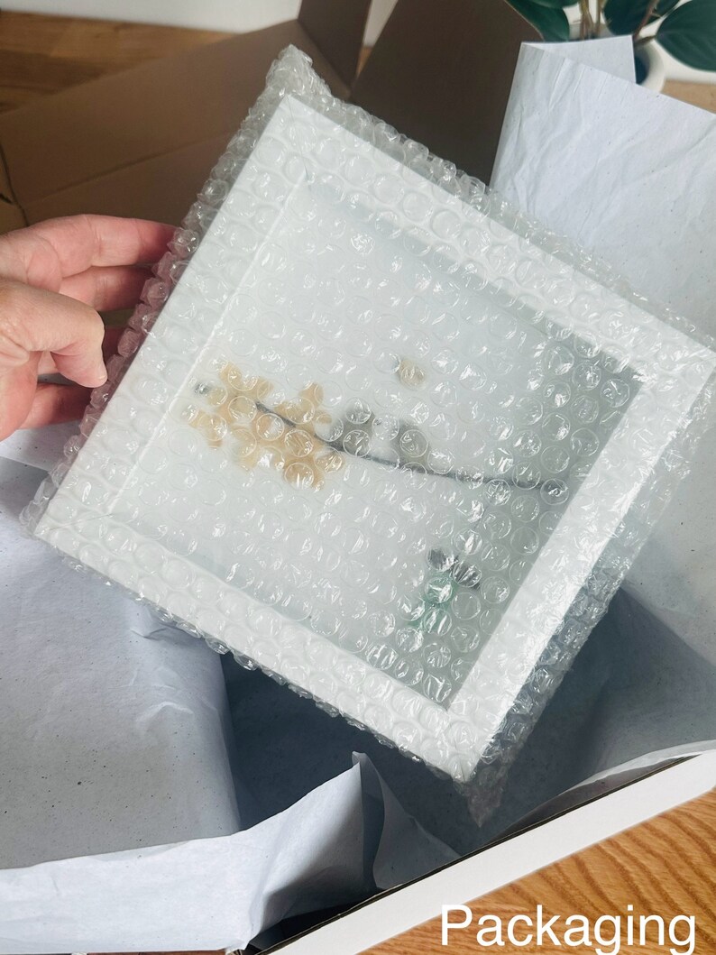 May include: A white frame with a clear plastic cover, protecting a delicate pressed flower arrangement. The frame is surrounded by bubble wrap for safe shipping.