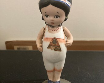 Puede incluir: Una muñeca de porcelana con cabello negro, un vestido blanco y un triángulo rojo y dorado en el frente que dice "Hiawatha". La muñeca está de pie sobre una base redonda gris.