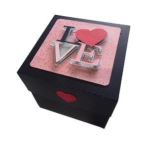 May include: A black gift box with a pink heart-patterned lid. The word "LOVE" is displayed on the lid in layered letters, with a red heart above the "E". A red heart is also on the front of the box.
