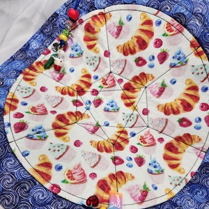 May include: A blue and white fabric circle with a variety of colorful cupcakes, croissants, and strawberries printed on it. The circle is surrounded by a blue fabric border with a swirling pattern. A colorful lollipop and a charm with the word "Sweet" are attached to the circle.