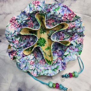 May include: A drawstring pouch with a floral print featuring succulents and roses in shades of pink, blue, and green. The pouch has a star-shaped interior with five compartments. A turquoise cord with beads and charms is attached to the pouch.