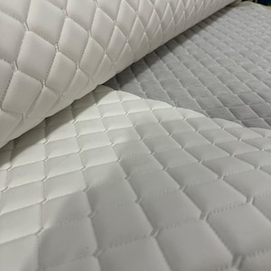 Pode incluir: Um rolo de tecido branco com um padrão de diamante acolchoado e detalhes de costura. O material tem uma textura lisa e almofadada, sendo mostrado parcialmente desenrolado, revelando a grade geométrica consistente na superfície do tecido.