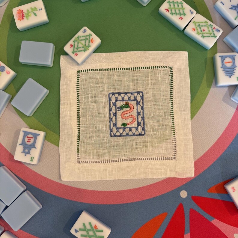 May include: A white linen square napkin with a green stitched border and a central embroidered design. The design features a blue frame with a pink and green snake. Mahjong tiles in various colors surround the napkin.