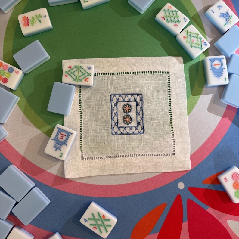 May include: A collection of Mahjong tiles and a white linen napkin with a blue embroidered design. The tiles are white with various colorful symbols and blue backs. The napkin has a stitched border.