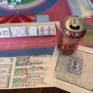 May include: A can of La Croix sparkling water sits on a white linen napkin with a blue and white embroidered design. Mahjong tiles and game instructions are visible on a pink and blue surface.
