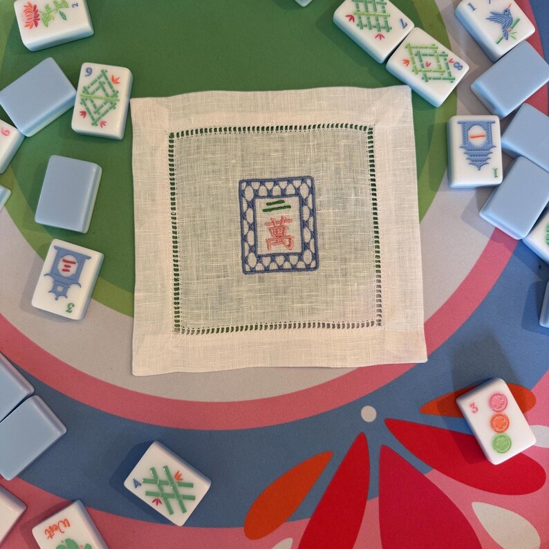 May include: A white linen coaster with a green stitched border and a blue and red embroidered design. Surrounding the coaster are various Mahjong tiles in blue, white, and green.