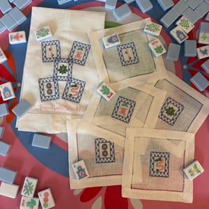 May include: A collection of white linen napkins and a matching towel, embroidered with blue and green designs, and surrounded by light blue Mahjong tiles. The towel has the text "BamDollCrack!" embroidered on it.