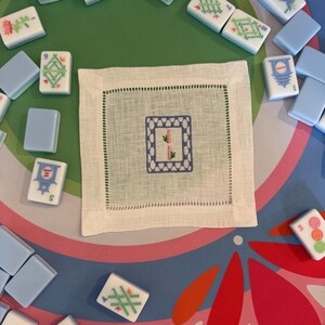 May include: A white linen square coaster with a blue embroidered bamboo design, surrounded by a green stitched border. Light blue and white Mahjong tiles are scattered around the coaster on a colorful floral patterned surface.
