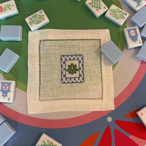 May include: A white linen square napkin with a blue and green embroidered floral design in the center. The napkin is surrounded by Mahjong tiles in various colors and designs, set on a green and patterned surface.
