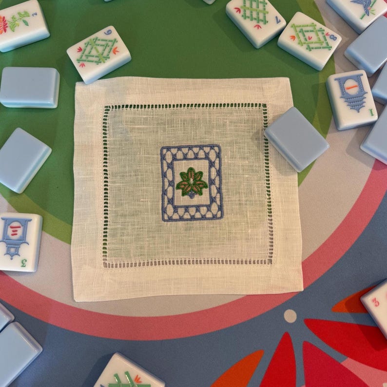 May include: A white linen square napkin with a blue and green embroidered floral design in the center. The napkin is surrounded by Mahjong tiles in various colors and designs, set on a green and patterned surface.