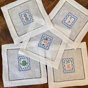 May include: Five white linen coasters with embroidered Mahjong tile designs. Each coaster features a different tile, framed by a blue stitched border and a black dotted edge. The coasters are square and rest on a wooden surface.