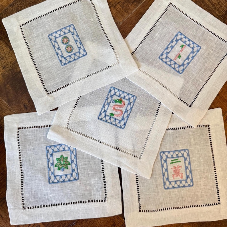 May include: Five white linen coasters with embroidered Mahjong tile designs. Each coaster features a different tile, framed by a blue stitched border and a black dotted edge. The coasters are square and rest on a wooden surface.