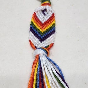 May include: A rainbow-colored friendship bracelet keychain with a white tassel. The keychain is made of colorful embroidery floss and has a silver metal key ring.