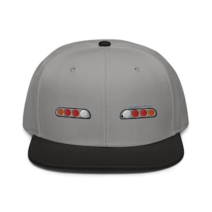 May include: A gray and black baseball cap with embroidered red, orange, and white taillights on each side. The text "Supra" is embroidered in white on the front of the cap.