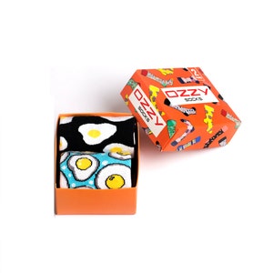 May include: A set of two pairs of socks in a bright orange box. The socks feature a fun, colourful design of fried eggs. The box has the text "OzZy Socks" printed on it.