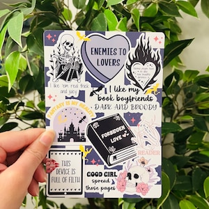 May include: A set of stickers with a dark and gothic aesthetic. The stickers feature a skeleton reading a book, a heart with the text "Enemies to Lovers", a book with the text "Forbidden Love", a castle with stars, a skull with flowers, and a tablet with the text "This device is full of filth".
