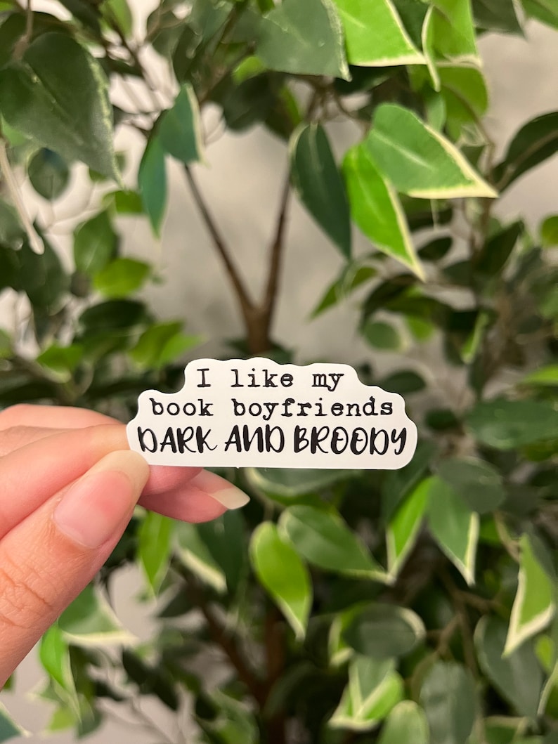 Op de afbeelding: Een witte sticker met zwarte tekst met de tekst "I like my book boyfriends DARK AND BROODY." De sticker wordt voor een achtergrond van groene bladeren gehouden.