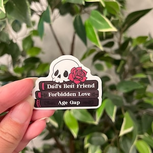 Op de afbeelding: Een sticker met een schedel met een rode roos en drie gestapelde boeken. De boeken zijn getiteld "Dad's Best Friend", "Forbidden Love" en "Age Gap". De sticker bevindt zich tegen een achtergrond van groene bladeren.
