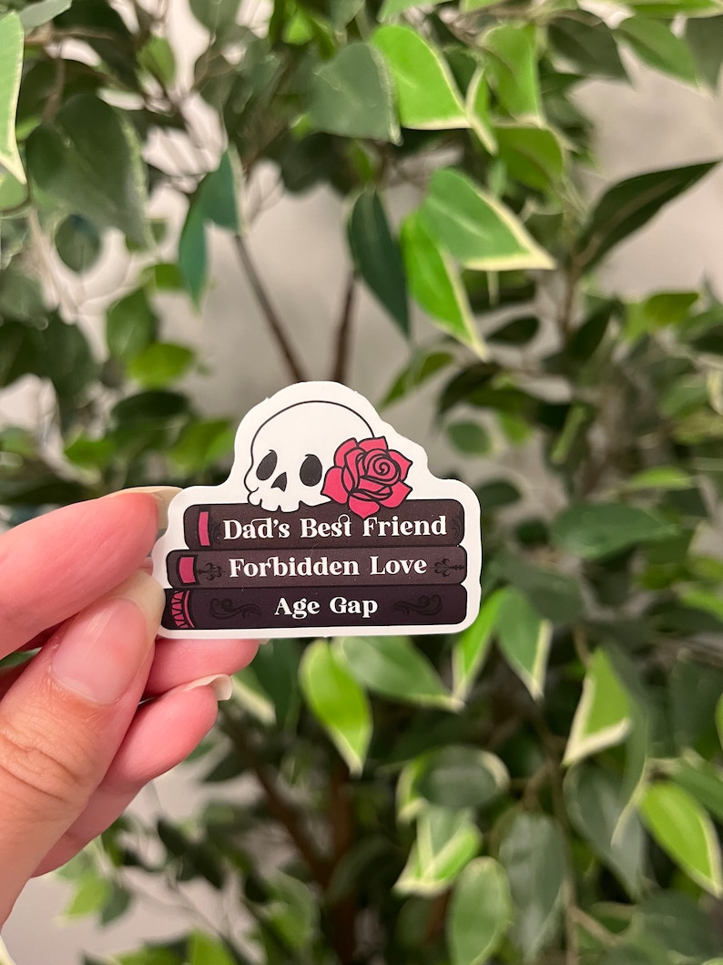 Op de afbeelding: Een sticker met een schedel met een rode roos en drie gestapelde boeken. De boeken zijn getiteld "Dad's Best Friend", "Forbidden Love" en "Age Gap". De sticker bevindt zich tegen een achtergrond van groene bladeren.