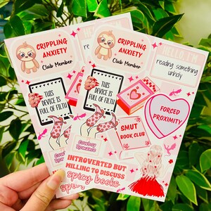 May include: A set of pink and red stickers with illustrations of sloths, books, and a woman in a red dress. The stickers feature text such as "Crippling Anxiety Club Member", "This device is full of filth", "Smut Book Club", "Introverted but willing to discuss spicy books", and "Forced Proximity".