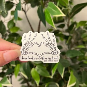 Op de afbeelding: Witte sticker met een zwart skelet hand hart ontwerp. De tekst luidt "I love books as dark as my heart" met "Dark Romance Addict" eronder. De sticker wordt getoond tegen een groene achtergrond.